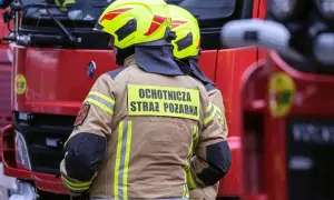 Strażacy w żółtych hełmach i beżowych kurtkach z napisem "Ochotnicza Straż Pożarna" stoją obok czerwonego wozu strażackiego.