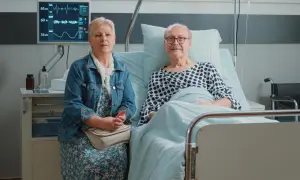 Starsza kobieta siedzi obok starszego mężczyzny w łóżku szpitalnym, na monitorze widoczne są parametry życiowe.
