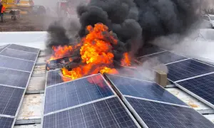 Płonące panele fotowoltaiczne na dachu, gęsty czarny dym i płomienie. W tle wozy strażackie i koparka. Wygląda na celowe podpalenie.