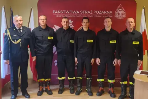 Sześciu strażaków w mundurach, w tym dowódca, tworzy rota strażacka. Stoją na tle sztandarów i logo Państwowej Straży Pożarnej.