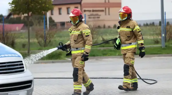 Dwóch strażaków w pełnym rynsztunku, jeden z wężem wodnym, ćwiczy akcję. Ciekawe, ile zarabia oficer straży pożarnej za taką pracę.