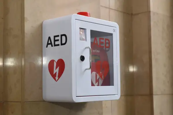 Biała szafka AED z czerwonym sercem i błyskawicą, zamontowana na ścianie.